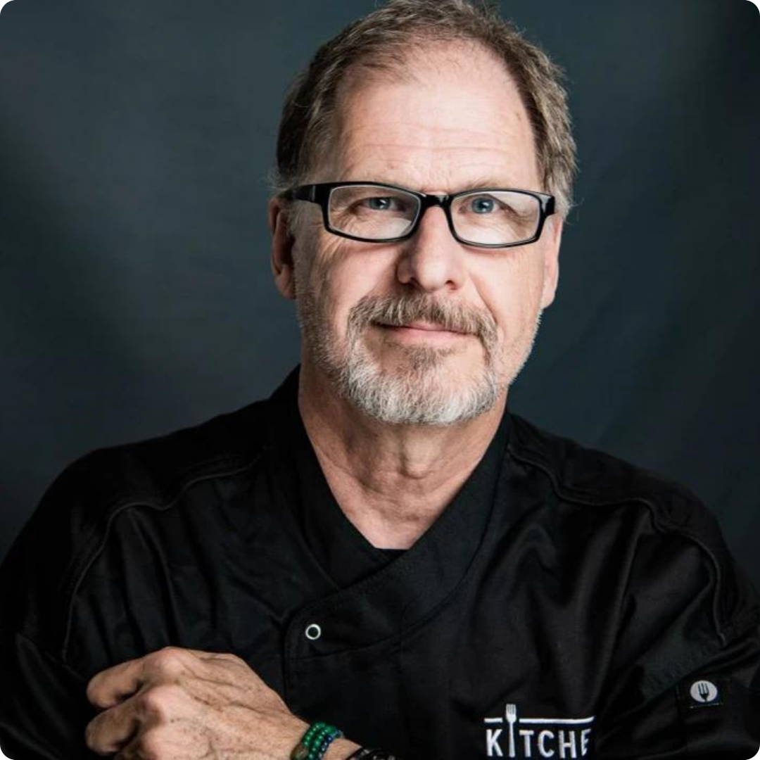 Chef with glasses, gray beard, black jacket, arms crossed, smiling.