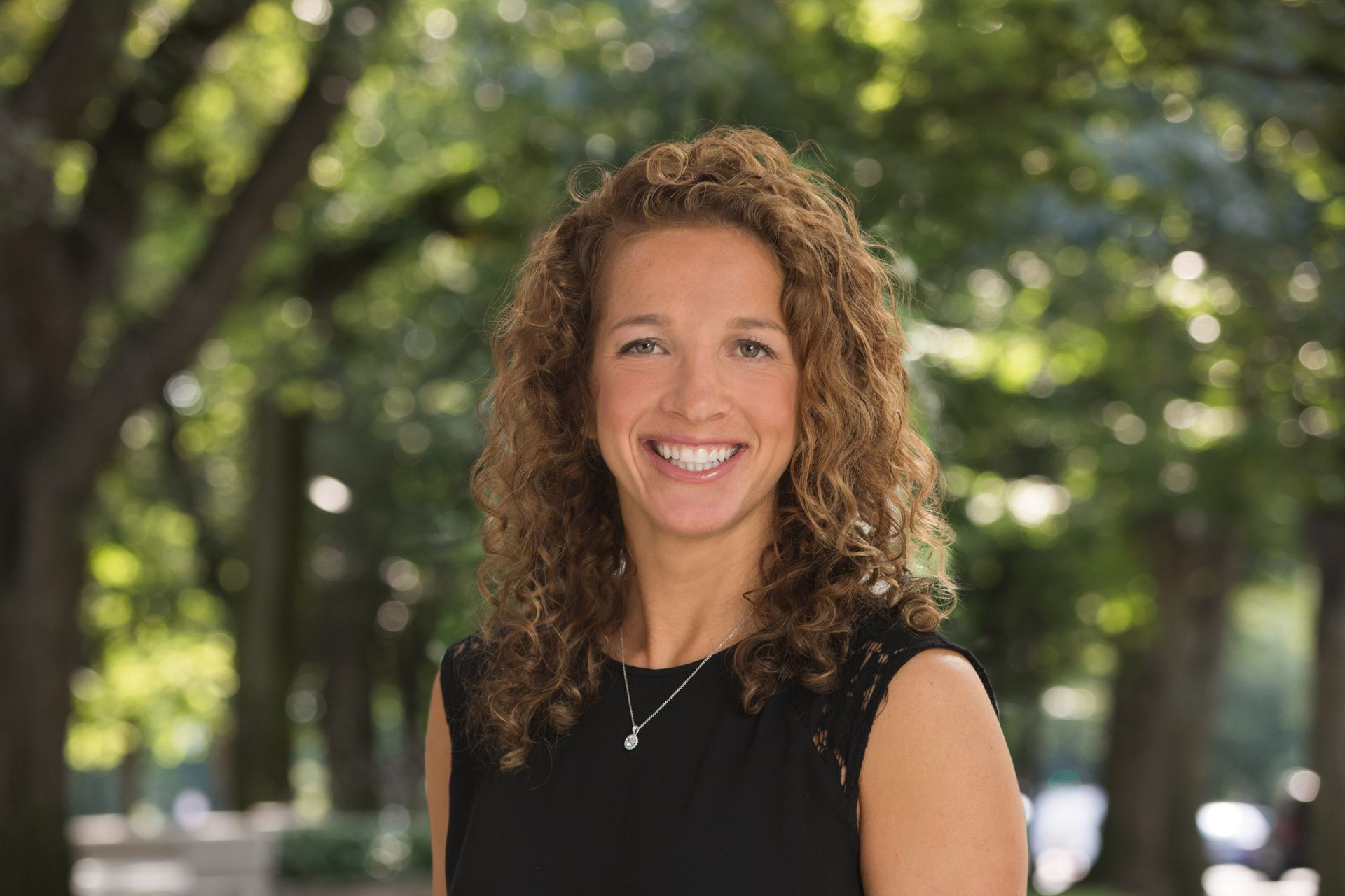 Woman with curly hair smiles outdoors, trees in background.