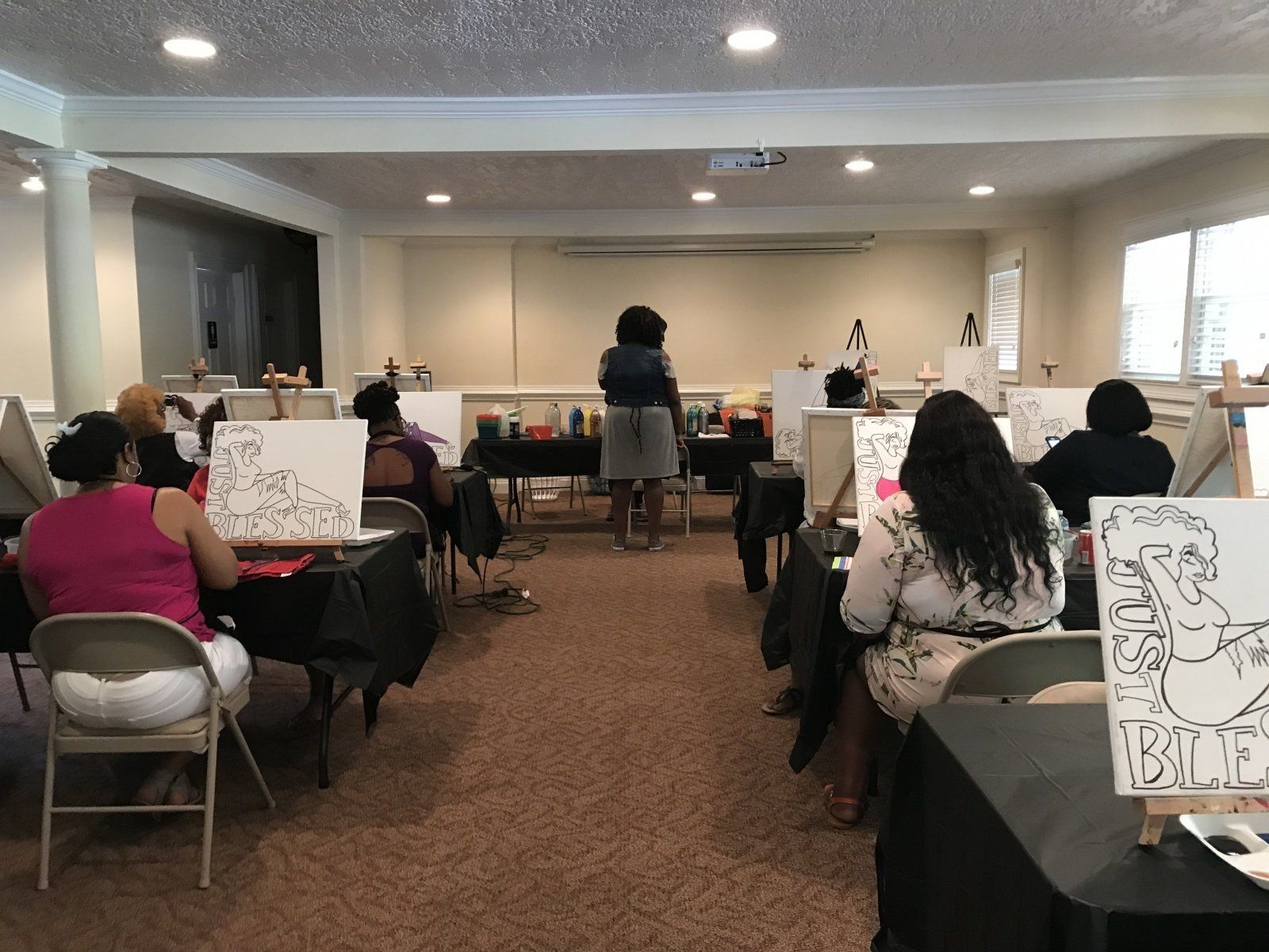 A group of people are sitting at tables in a room painting on canvases.