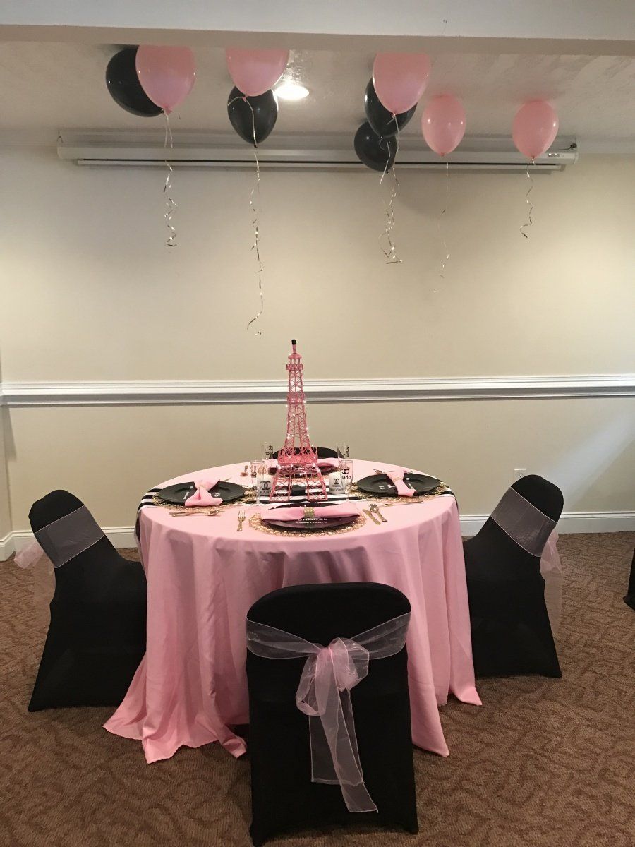 A table with a pink tablecloth and black chairs with balloons hanging from the ceiling.