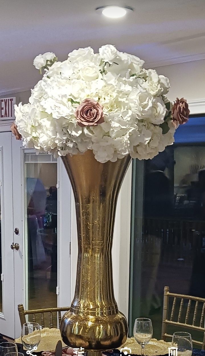 A tall gold vase filled with white flowers is on a table.