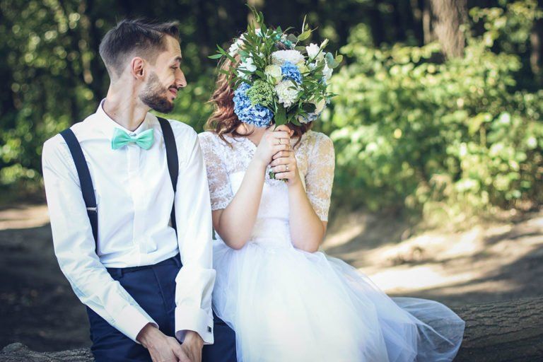 A bride and groom are sitting next to each other in the woods holding a bouquet of flowers.