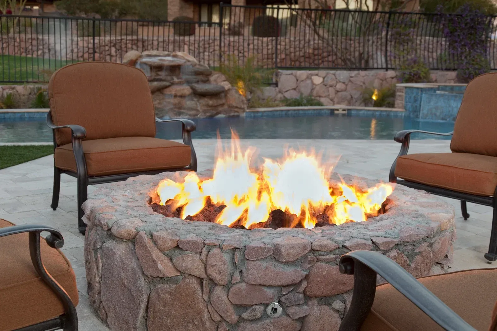 A fire pit is surrounded by chairs and a pool.