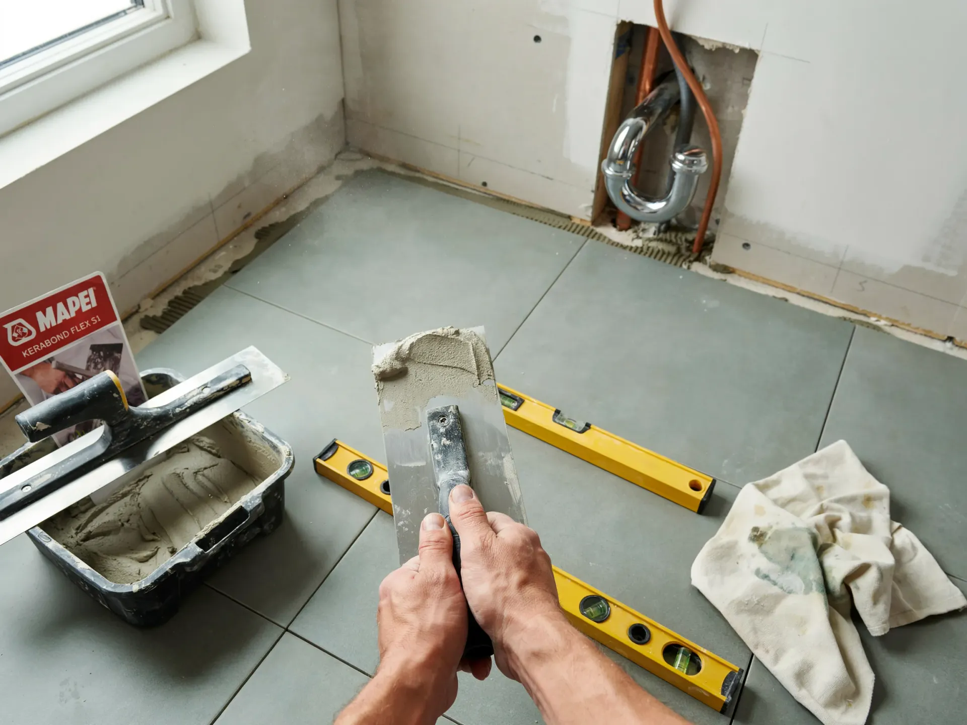 Hands holding a trowel over tiled floor in a bathroom renovation with level, bucket, and wall plumbing opening.