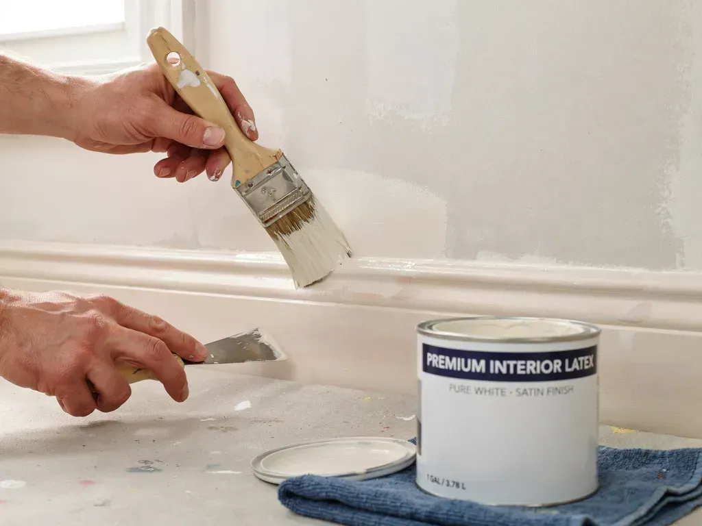 Hands painting white trim with a brush beside a paint can on the floor