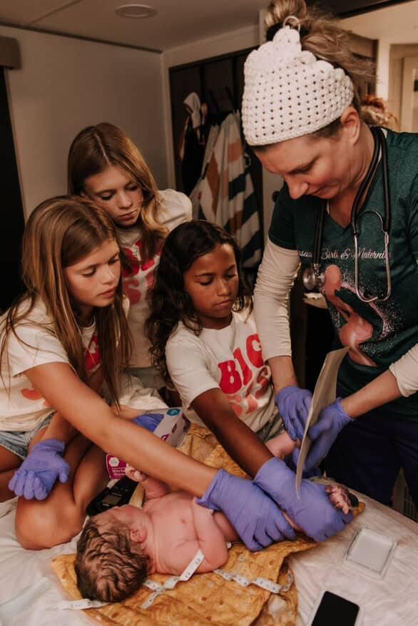 A newborn baby is examined by a midwife while three children look on.
