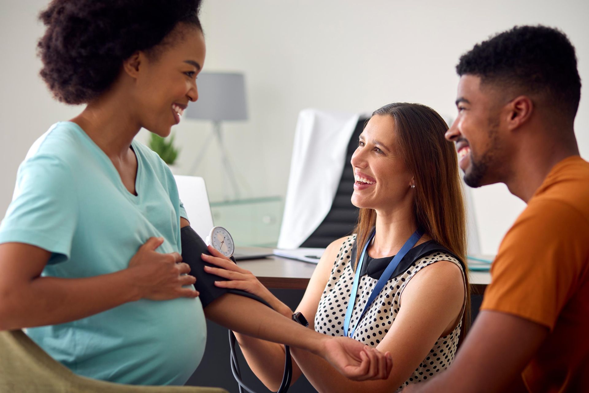 Pregnant woman has blood pressure checked by a medical professional, with her partner present. All are smiling.