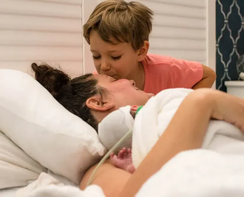 Mom in bed smiles as older child kisses her forehead; newborn visible.