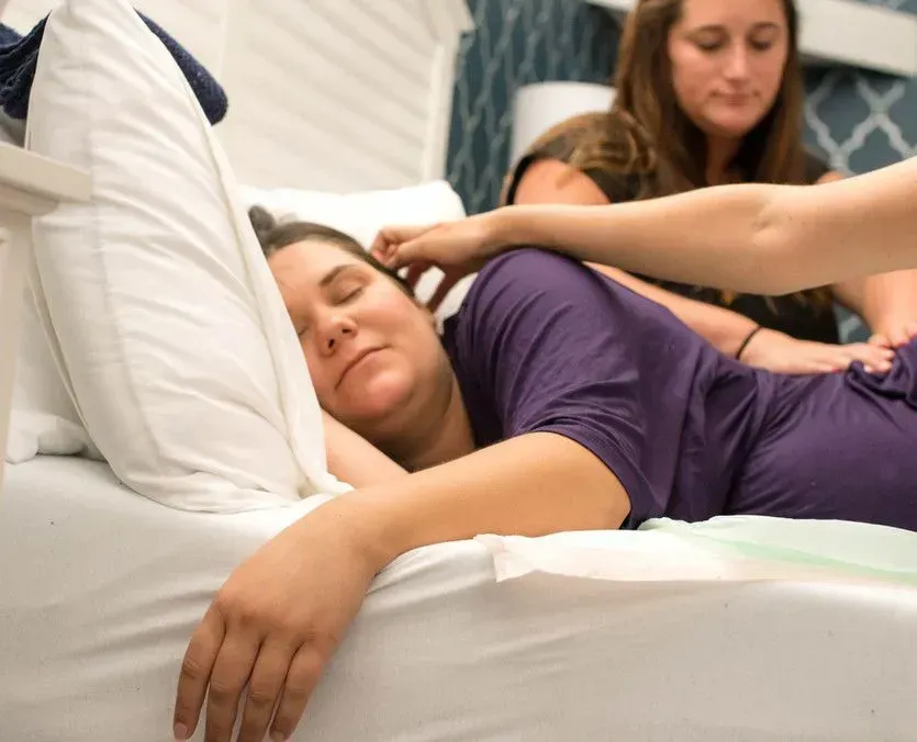 Woman resting in bed with hands on her head and back; another woman standing over her.