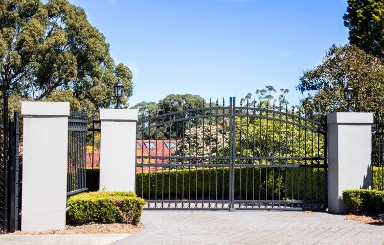 Black wrought-iron gate between two white pillars, driveway, green hedge and trees against a blue sky.