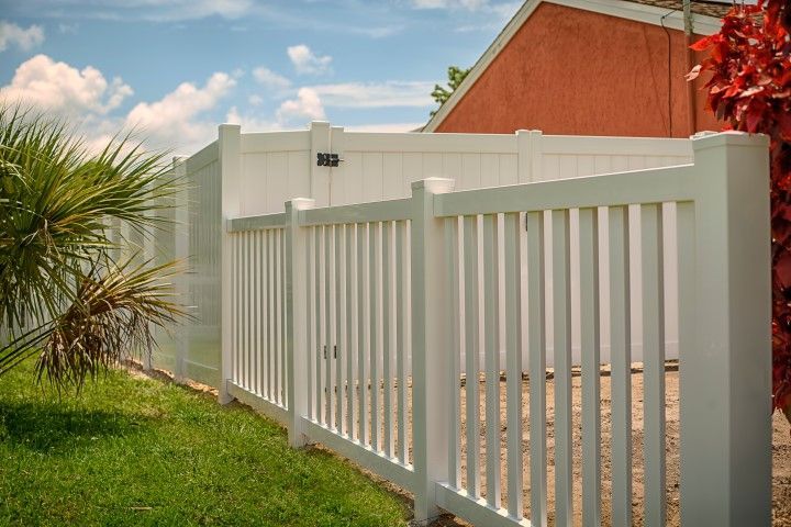 White vinyl picket fence surrounding a grassy yard with blue sky and palm tree in the background.