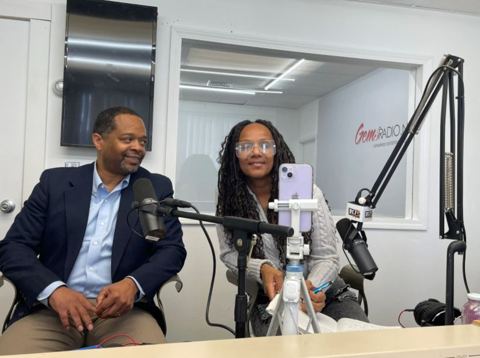 Two people in a radio studio: one man, one woman. They sit at a table with microphones, a phone on a tripod, and look toward the camera.