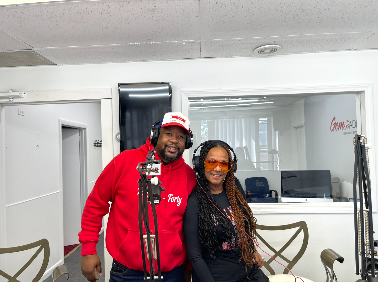 Two people in a recording studio. A man in red hoodie smiles, and a woman wears orange glasses and smiles. Microphones are present.
