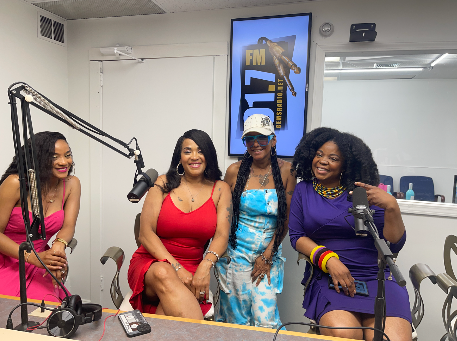 Four women at a radio station; two wearing red and purple dresses, and two with blue tops, smiling near microphones.