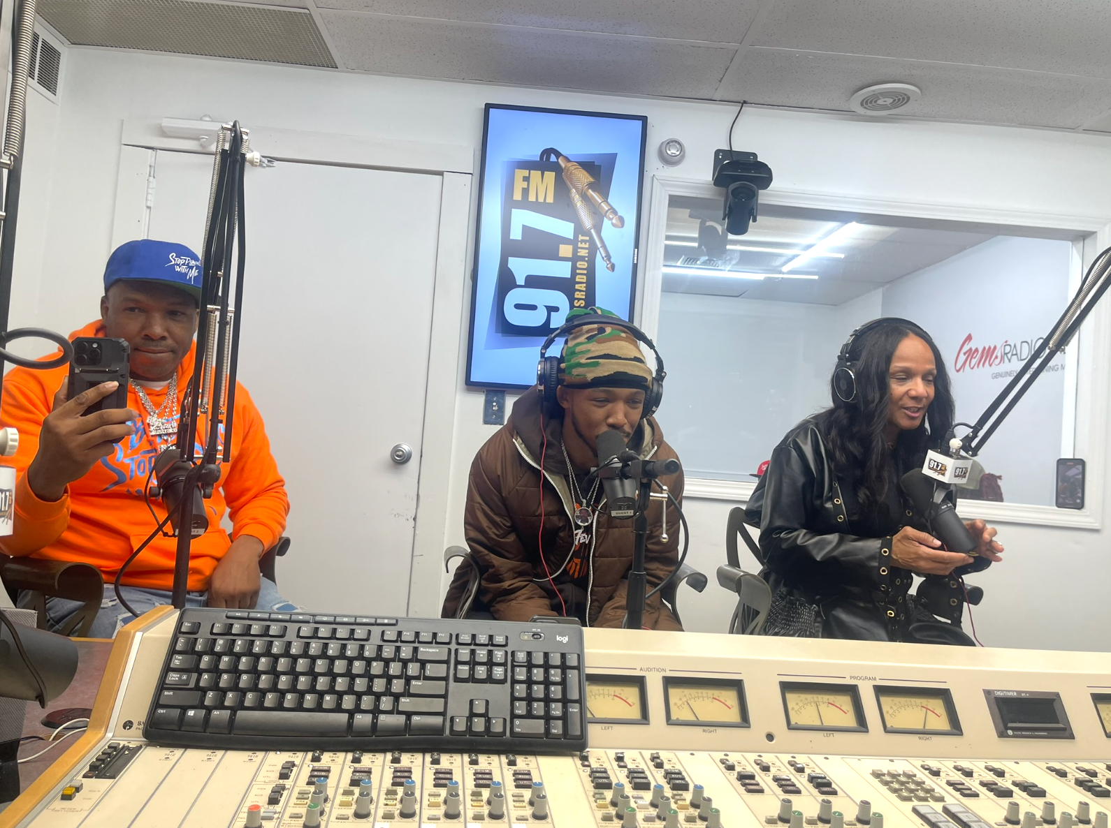 Three people in a radio studio at 91.7 FM. One holds phone, one speaks into mic, and one smiles at mic.