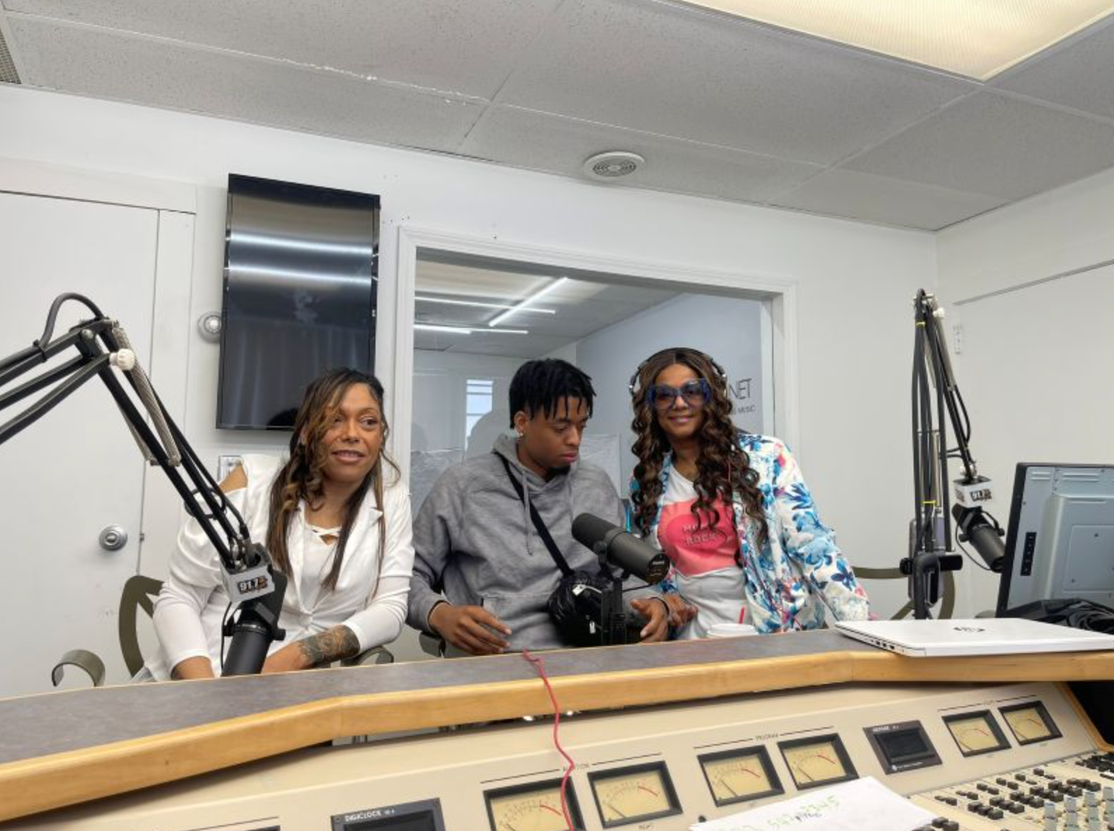 Three people at a radio station; two women and one person in a grey hoodie seated at a soundboard with microphones.