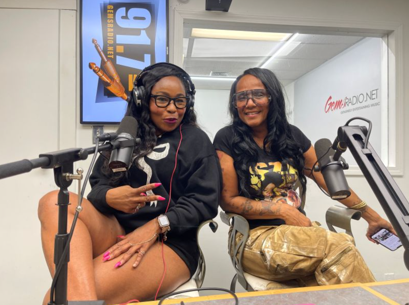 Two people in a radio studio, sitting at mics. One gestures peace sign; the other holds a phone. 
