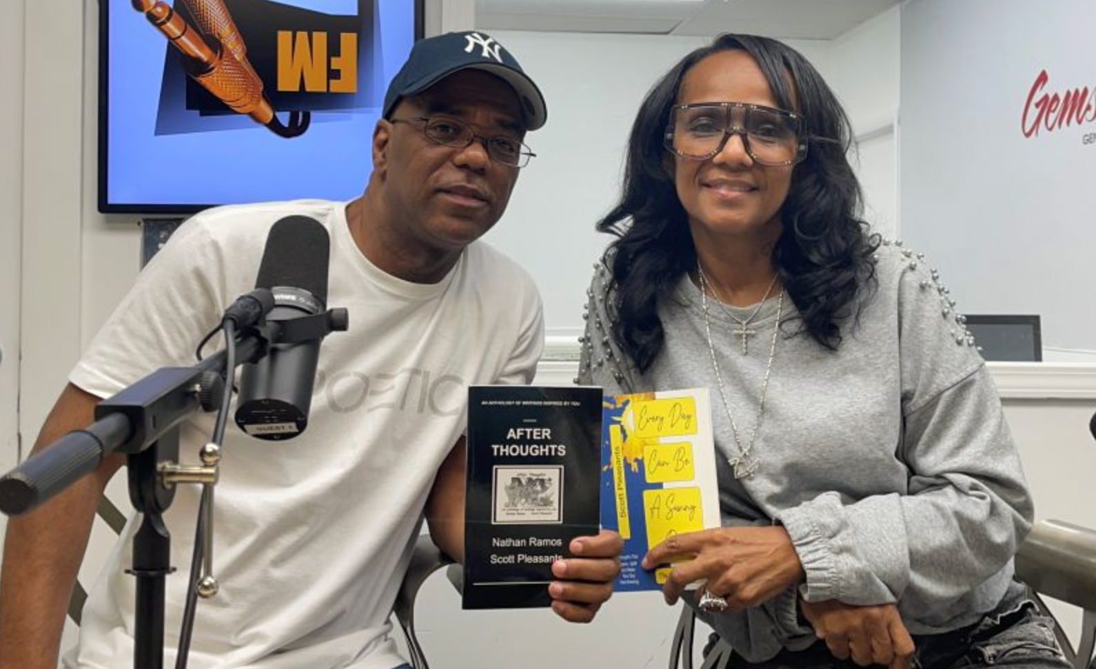 Two people holding books at a radio station. Man wears a cap and white t-shirt, woman wears glasses and gray sweater.