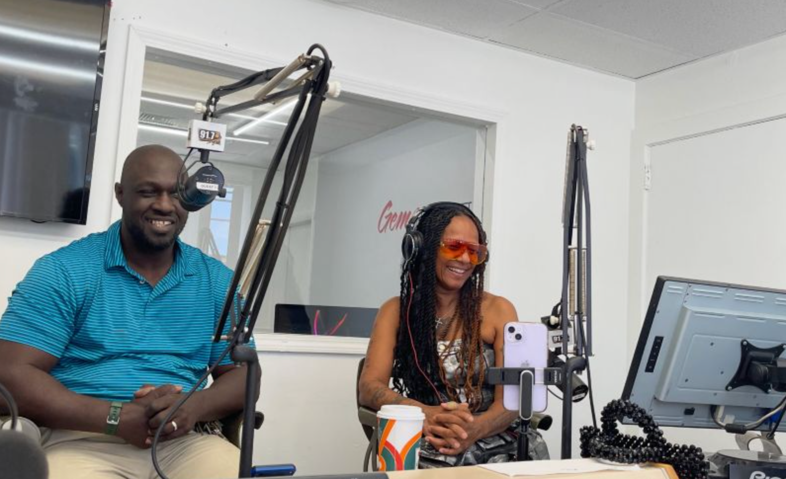 Two people in a radio studio; man smiles at mic, woman wears orange glasses.