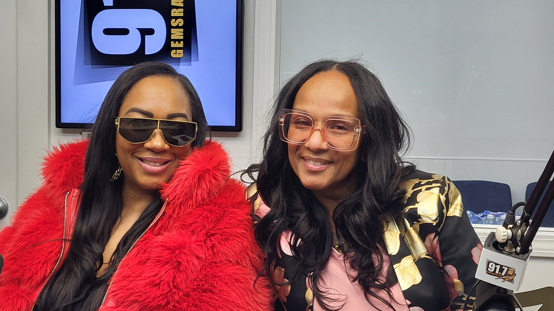 Two women smiling at a radio studio. One wears a red fluffy jacket and dark sunglasses, the other has patterned glasses and a floral top.