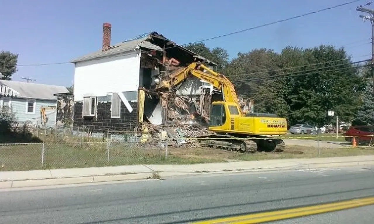 A yellow excavator is demolishing a house on the side of the road.