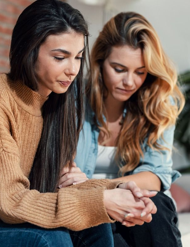 Two women with concerned expressions, one comforting the other by holding her hands.