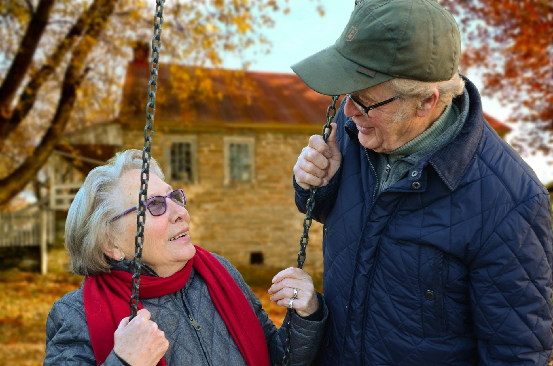Elderly couple on a swing set, smiling at each other in front of a house. Fall foliage surrounds them.