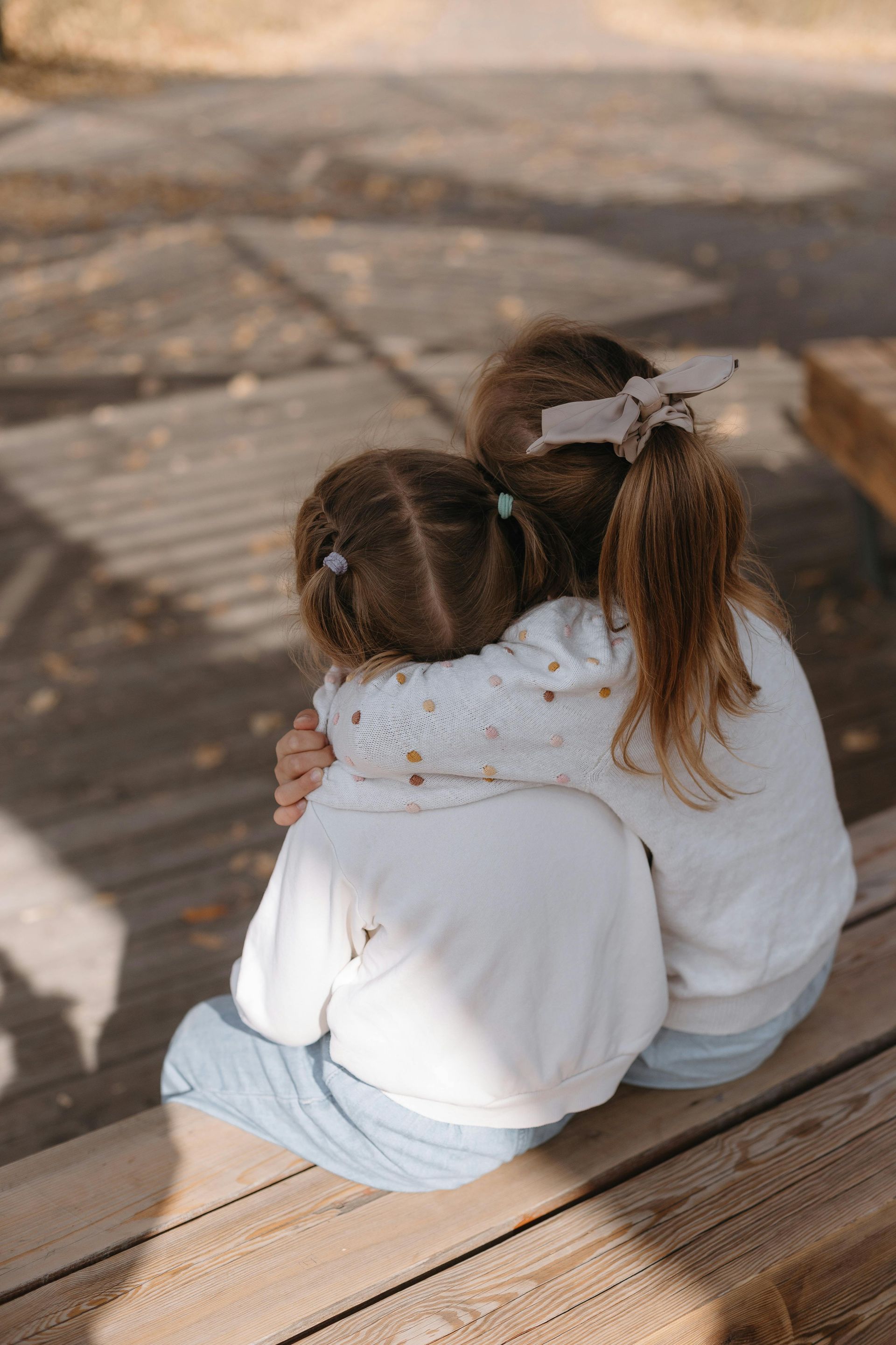 Two children, backs to camera, hugging on a wooden bench outdoors.