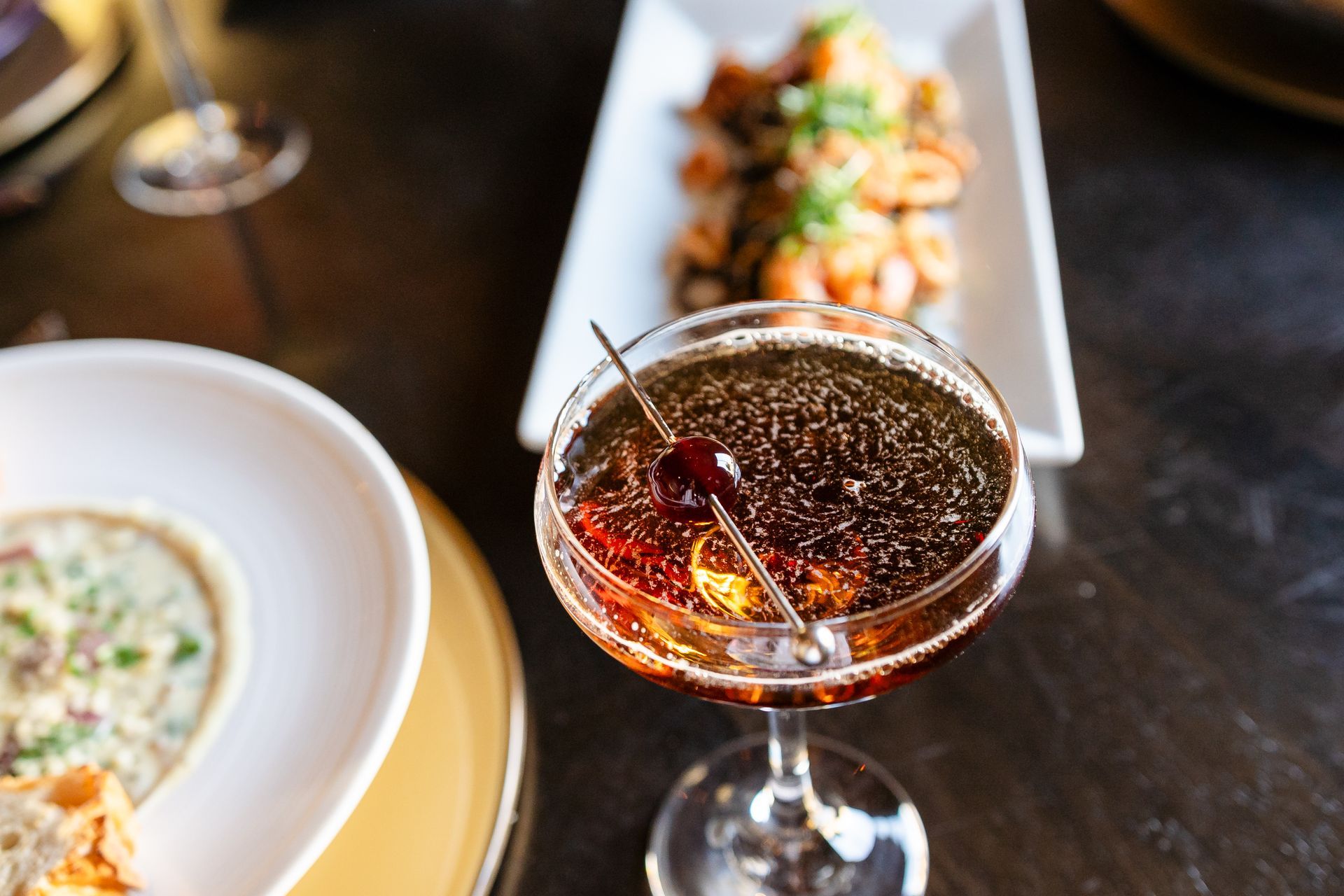 A martini with a cherry in it is on a table next to a plate of food.