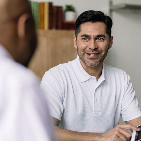 A man in a white shirt is sitting at a table talking to another man.