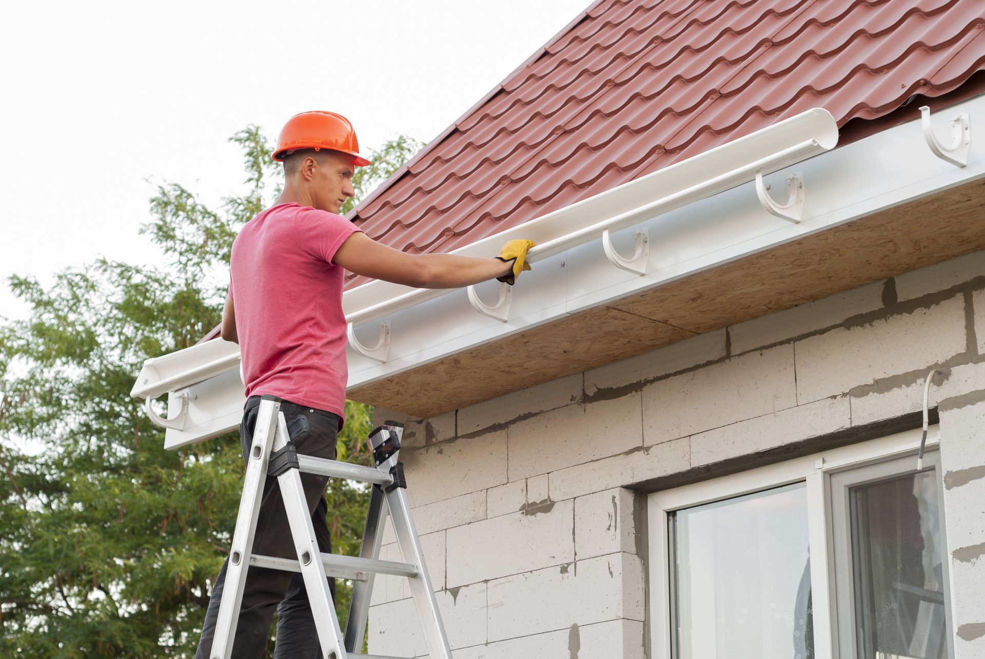 A man is standing on a ladder fixing a gutter on the side of a house.