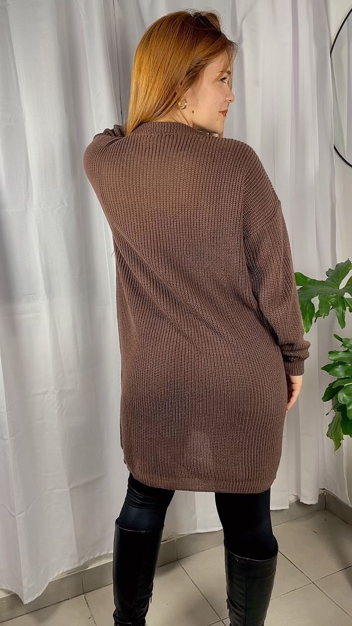 Mujer con un vestido de punto texturizado marrón y botas negras de pie en el interior de una casa junto a una cortina blanca.