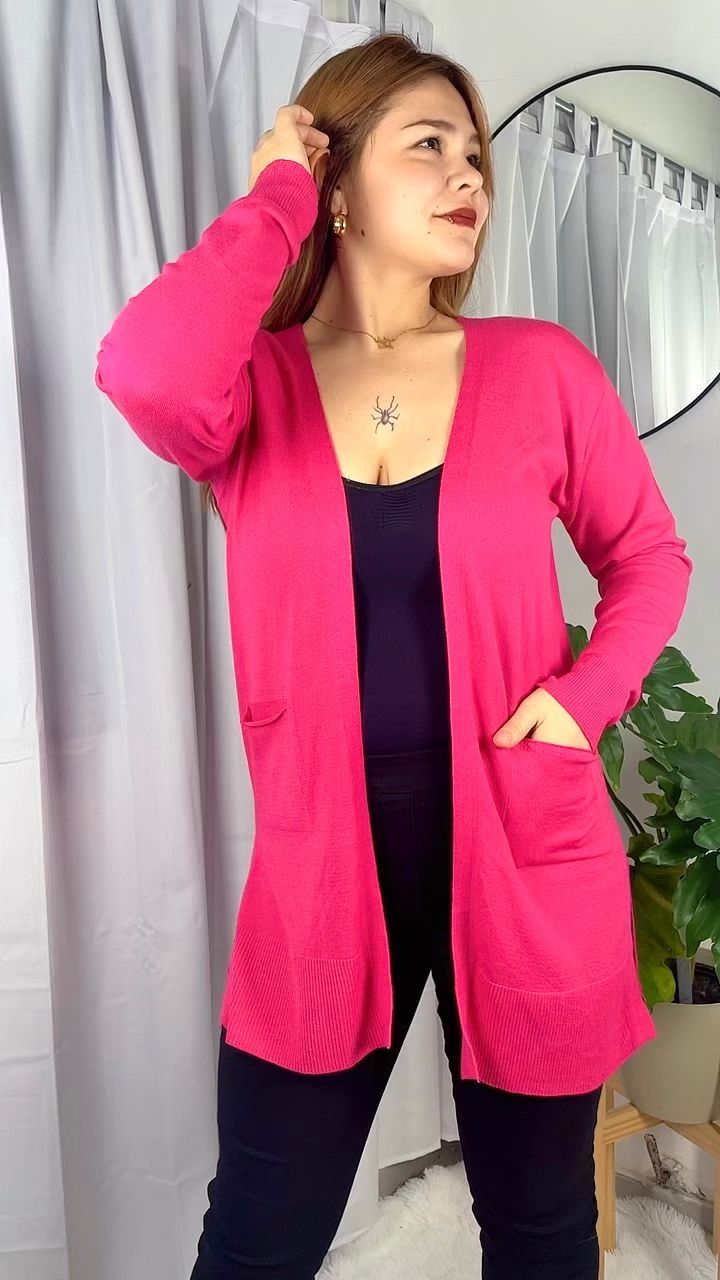 Mujer con un cárdigan rosa brillante sobre ropa negra, posando en el interior junto a unas cortinas y un espejo.
