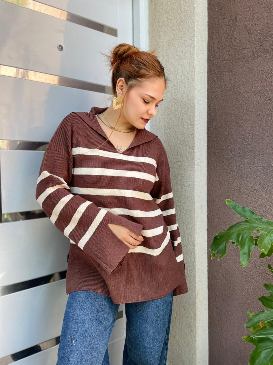 Mujer con suéter de rayas marrones y jeans de pie junto a una puerta al aire libre, mirando a la cámara.
