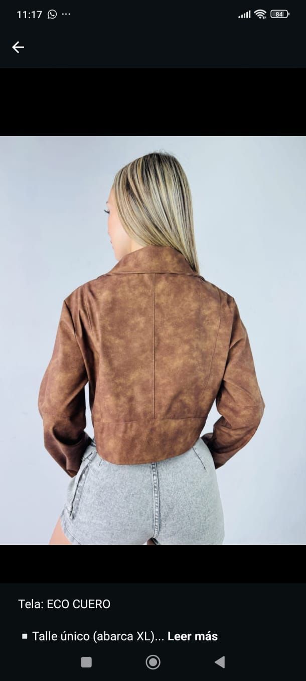 Mujer rubia con chaqueta marrón y falda gris posando frente a un fondo gris claro.