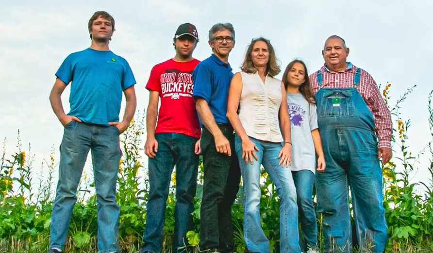 Six people stand together outdoors; includes a man in overalls and a woman in a light top, blue sky visible.
