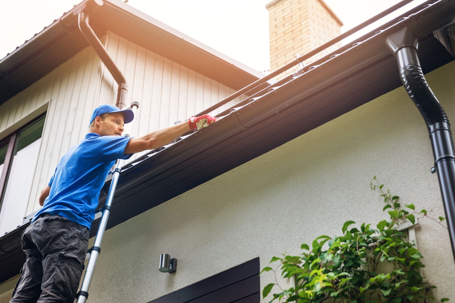 A person in a blue shirt standing on a ladder and cleaning out debris from a residential gutter.