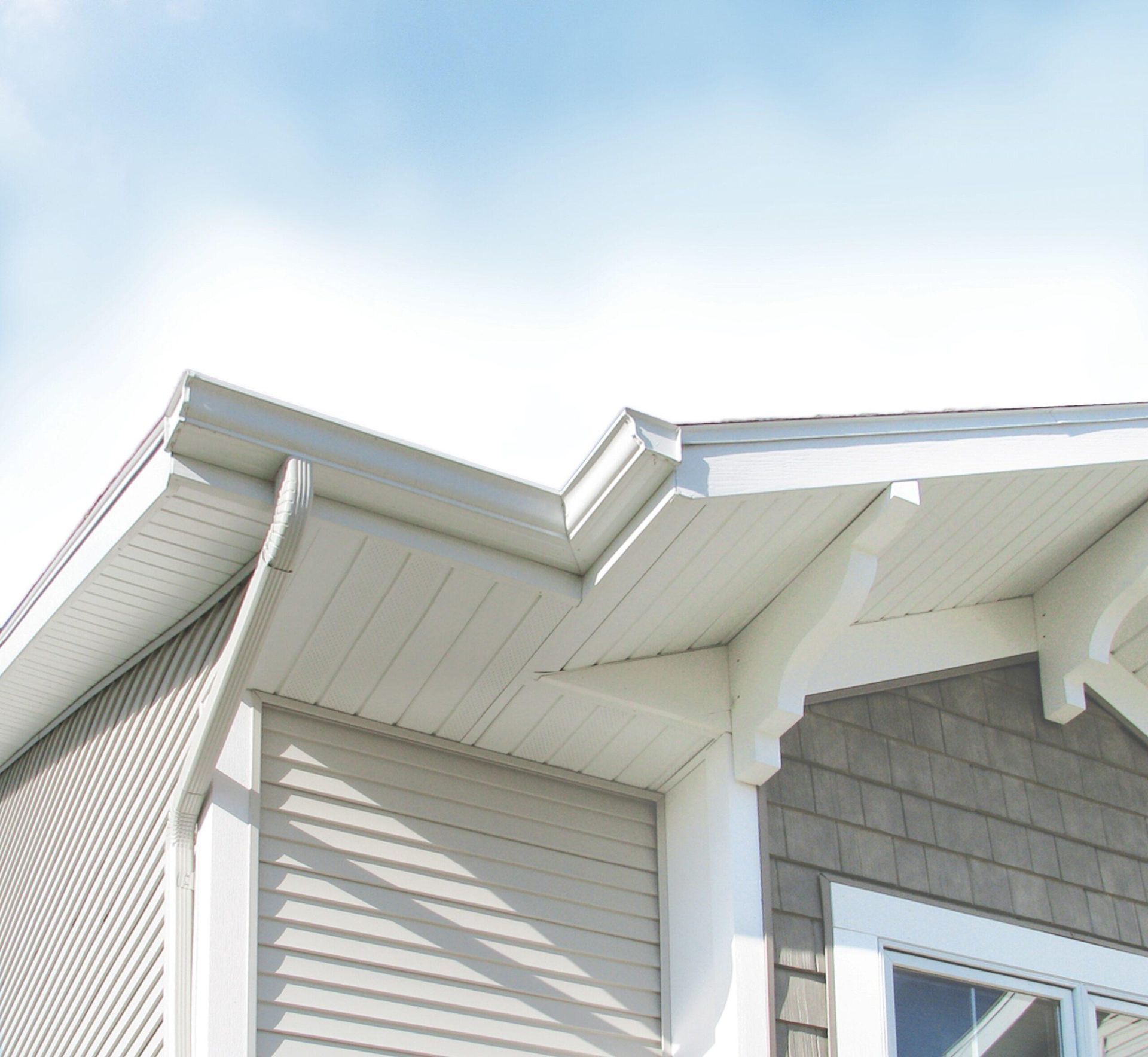 Low-angle shot of a tan-colored house exterior, showing roof eaves, soffits, and a downspout against a blue sky.