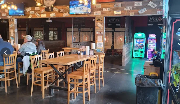 Interior of a bar with tables, chairs, TVs, arcade games, and dart boards. Customers sit at tables.