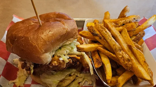 Chicken sandwich with coleslaw and lettuce, served with a side of fries on a red and white checkered paper.