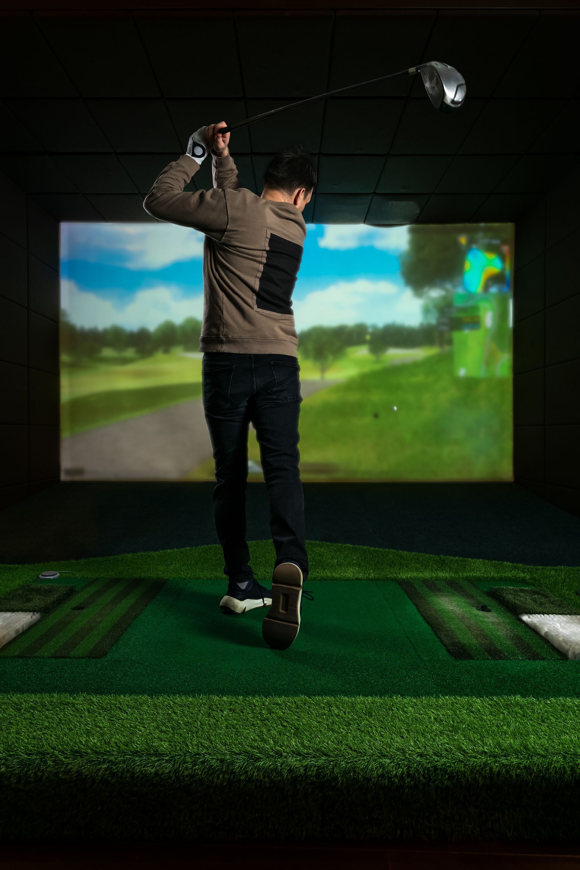 Man swings golf club in indoor golf simulator; green mat, screen displays golf course.