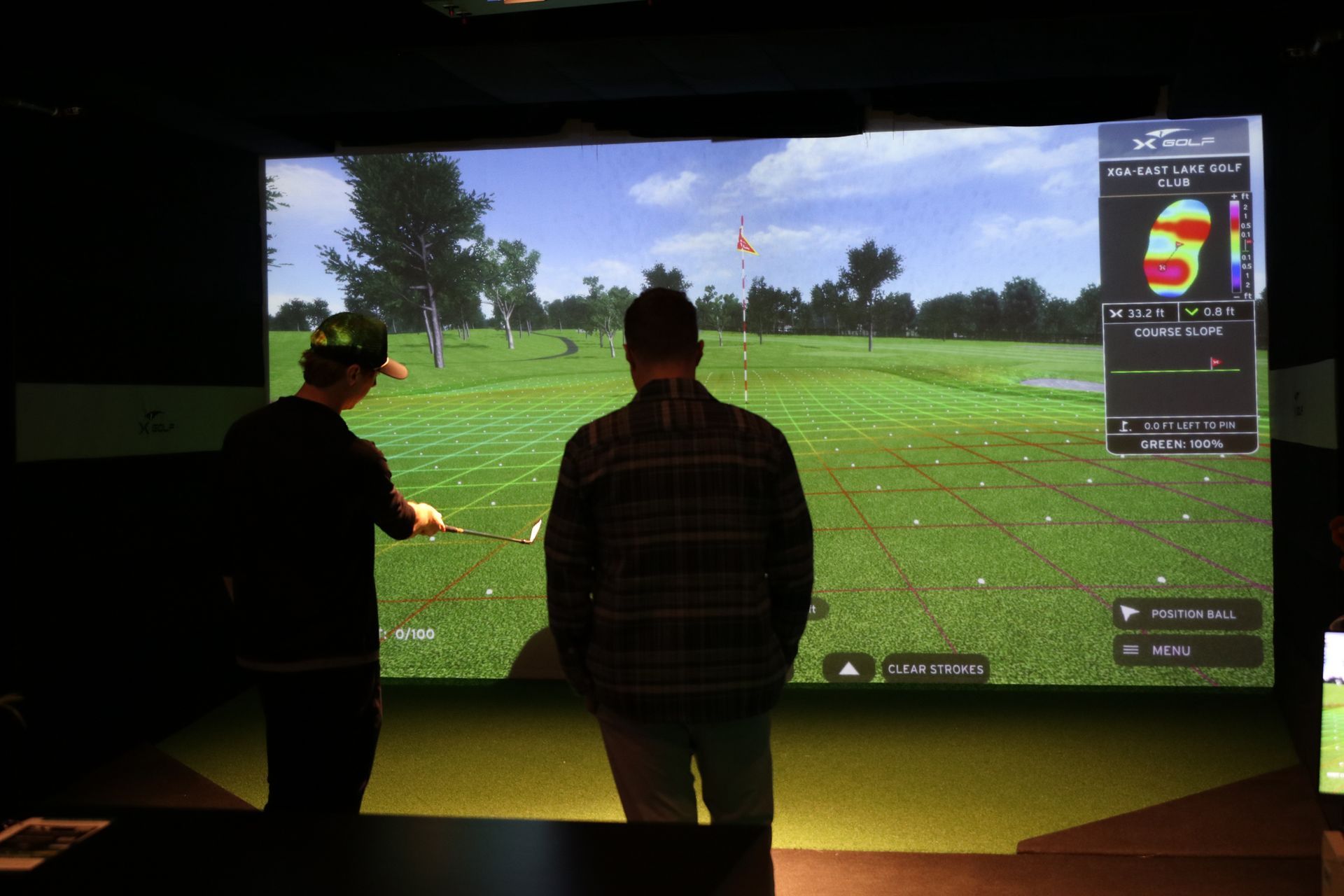 Two men at a golf simulator, looking at a screen displaying a course; one holds a golf club.