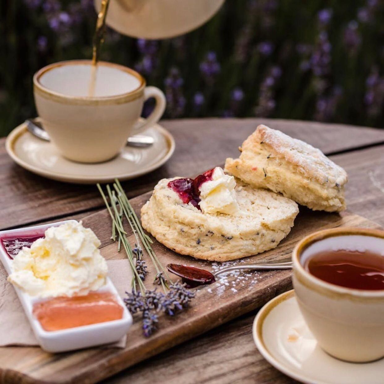 Tea being poured into cup next to scones with cream and jam on a wooden board outdoors.