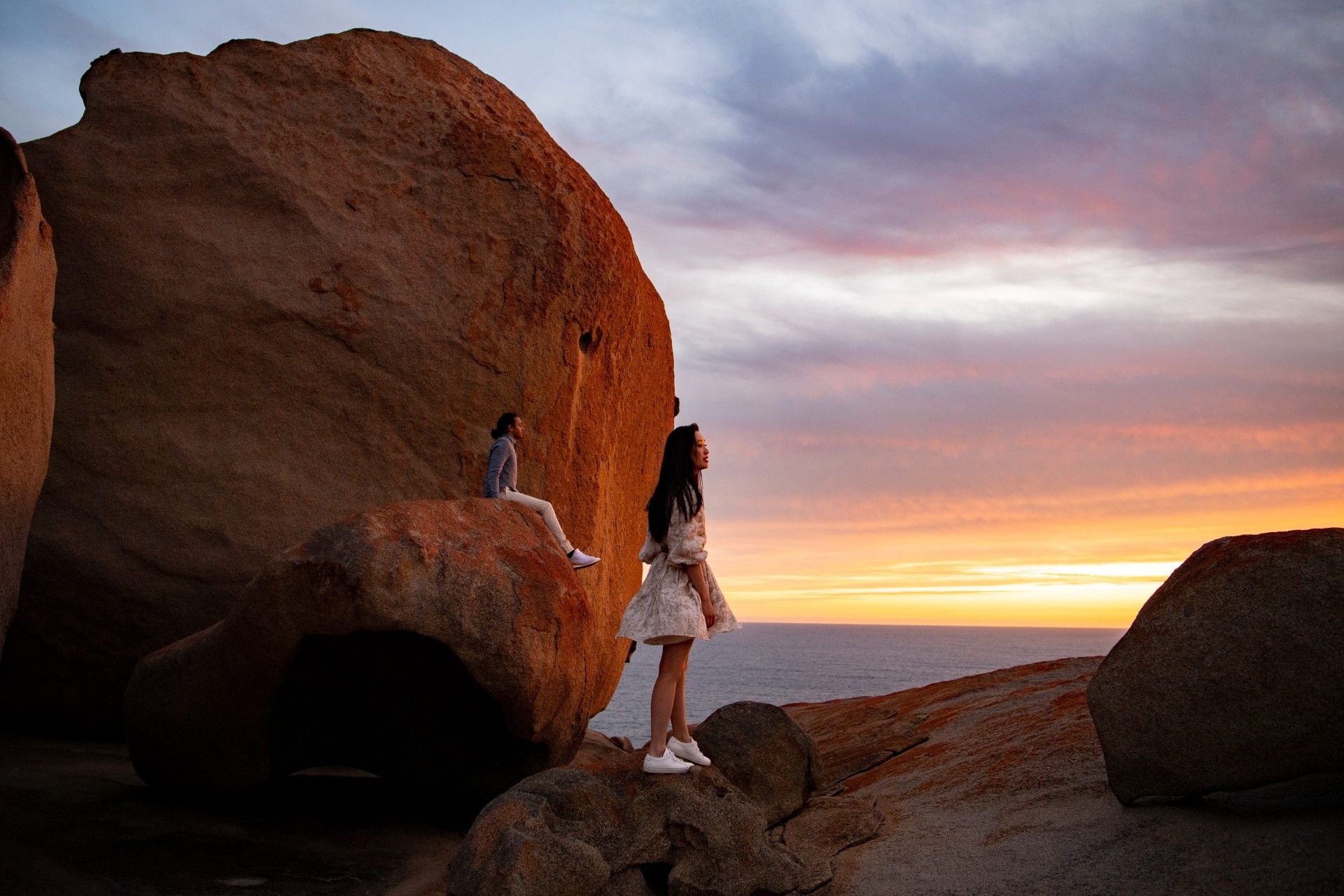 Two women on rocks overlooking the ocean at sunset. One standing, one sitting. Orange sky.