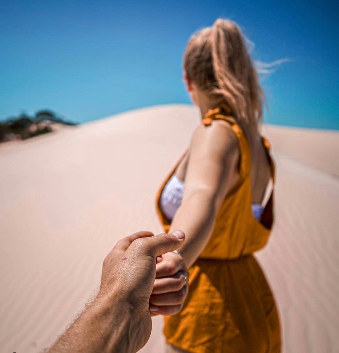 Woman in yellow dress leading someone's hand, walking on a sand dune under a blue sky