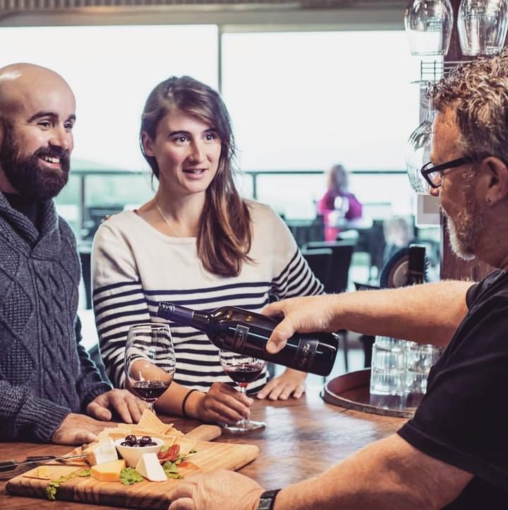 A bartender pours red wine for a couple at a wooden bar