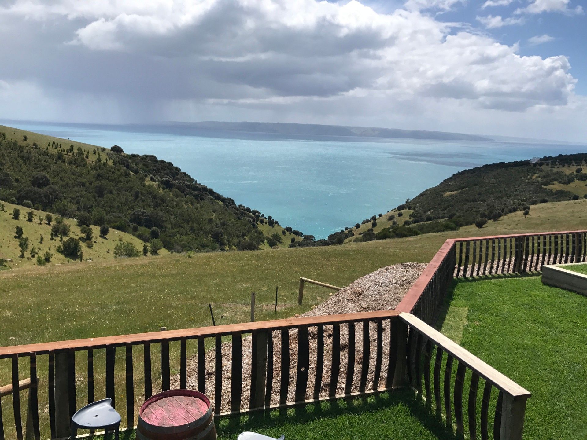 Rolling green hills overlooking turquoise water, viewed from a wooden deck with a barrel and grass.