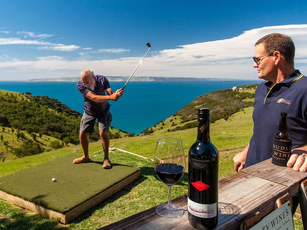 Man swings golf club off a hill overlooking ocean, another man watches, wine bottle and glass on rail.