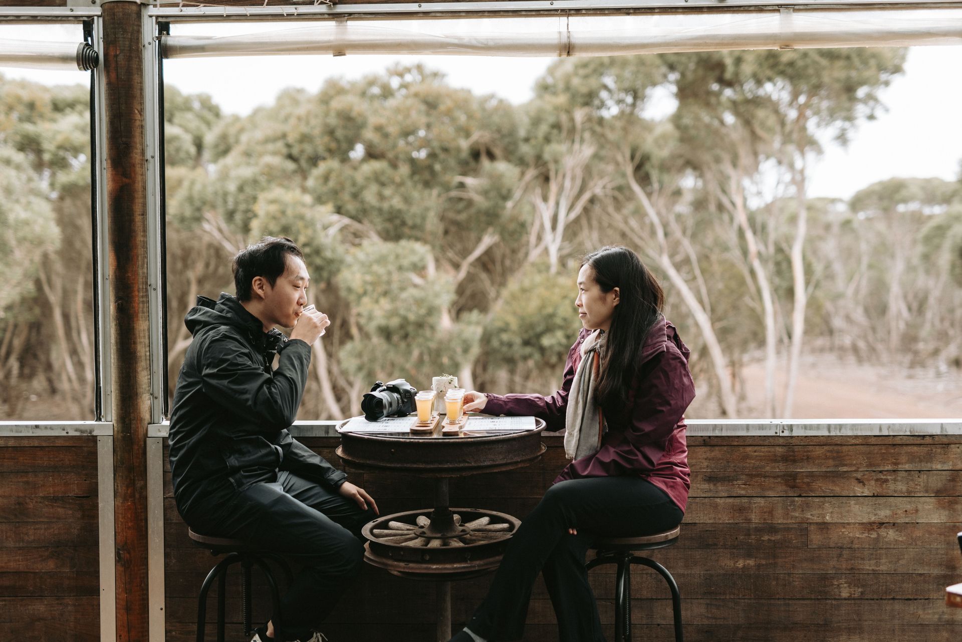Couple at outdoor bar, trees in background. Man sipping drink, woman talking.