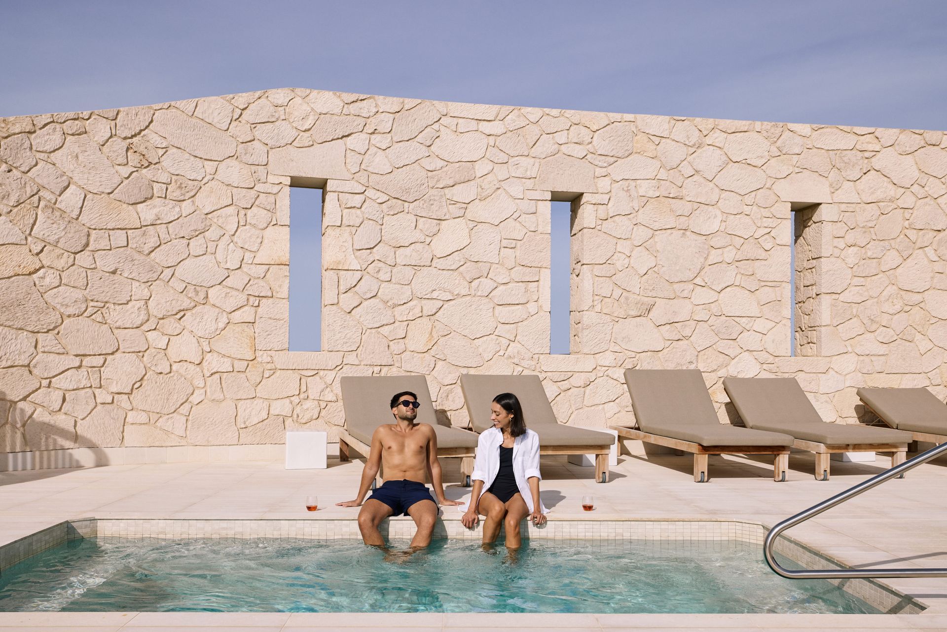 Couple sits by pool with feet in the water, against a stone wall with three windows and lounge chairs.